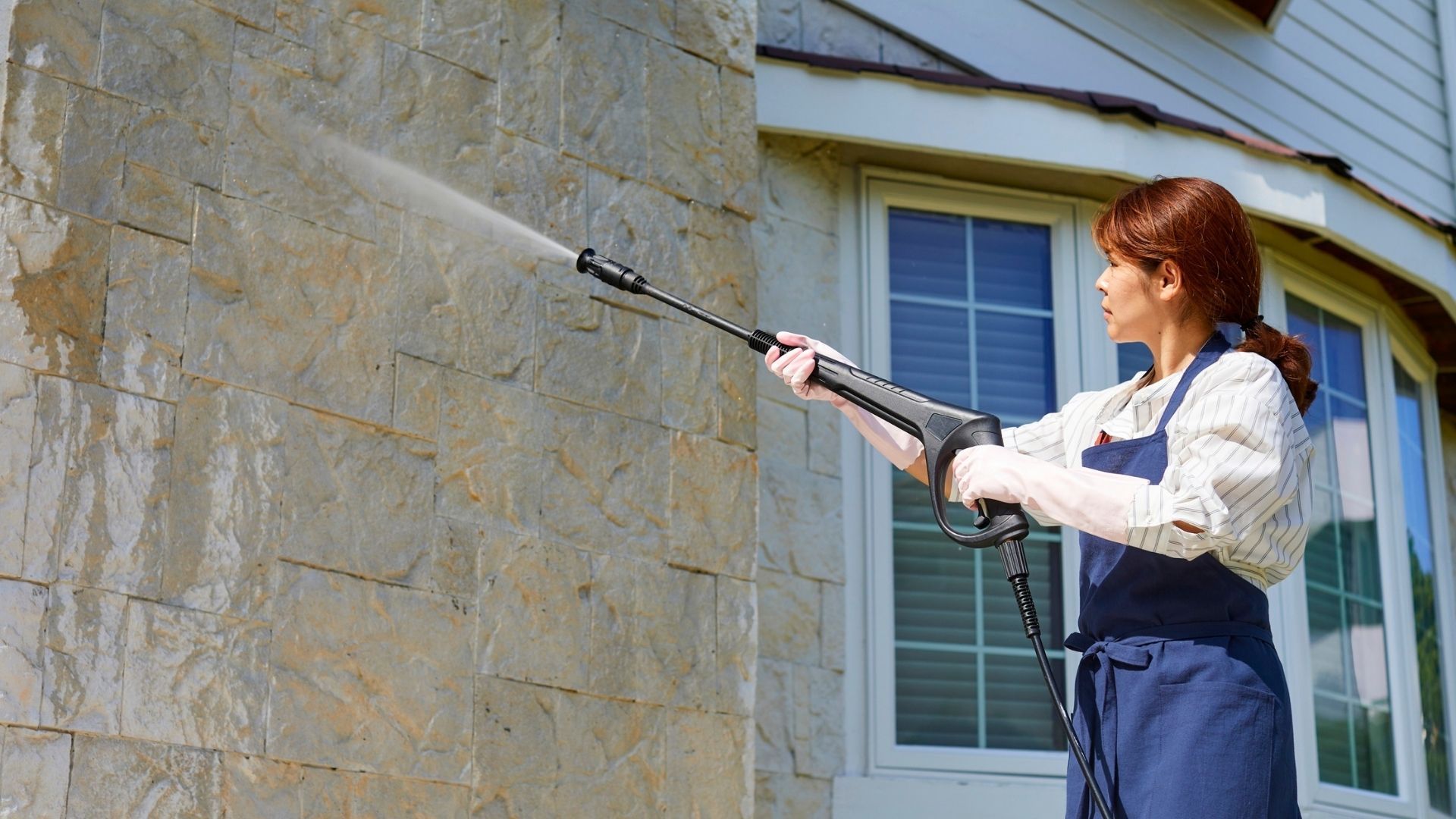 Woman power washing a stone house exterior near a window
