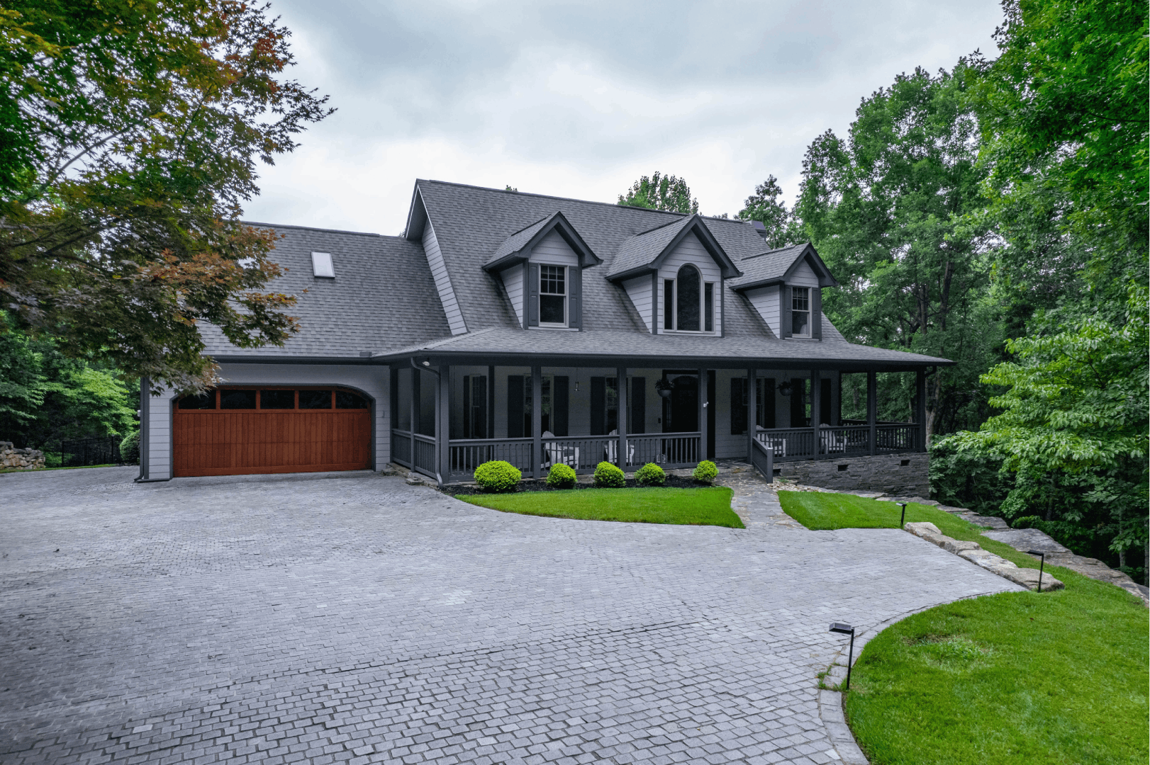Residential exterior: Gray house with a large porch and a wood garage door surrounded by trees.