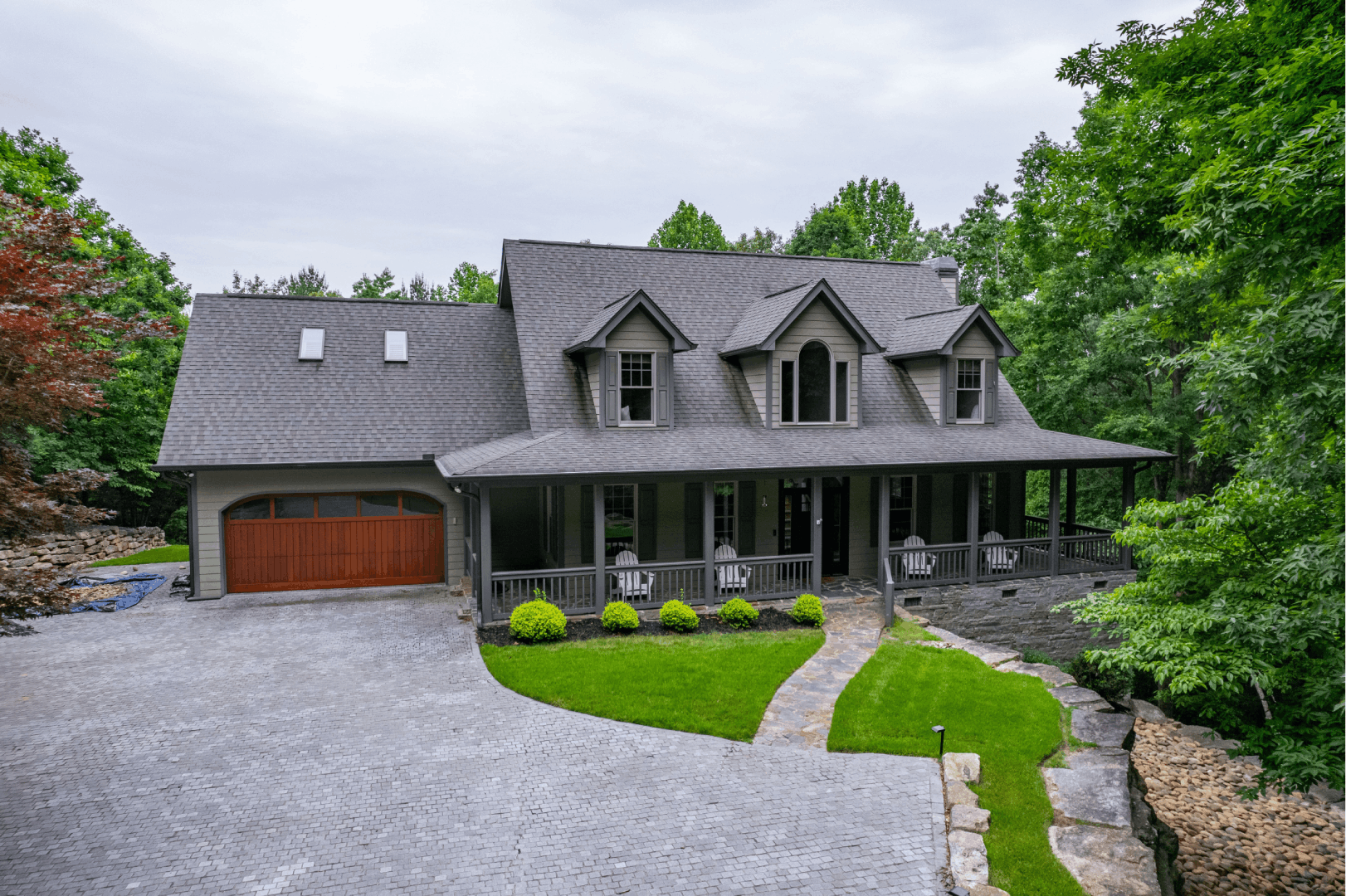 Residential exterior: Gray house with a large wrap-around porch, surrounded by lush green trees.