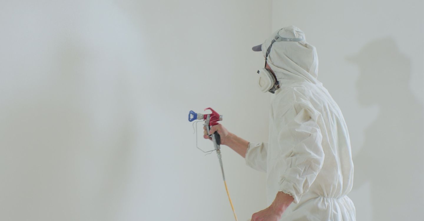 Painter in protective gear spraying a white wall with a paint sprayer.