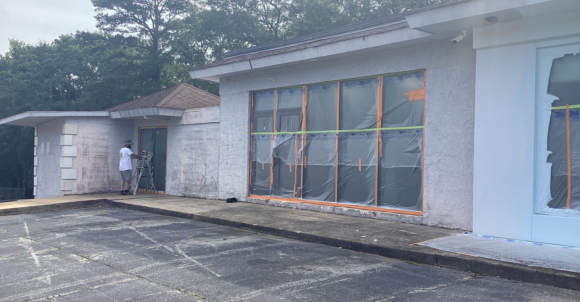 Commercial building exterior painting prep: windows covered, worker on ladder.