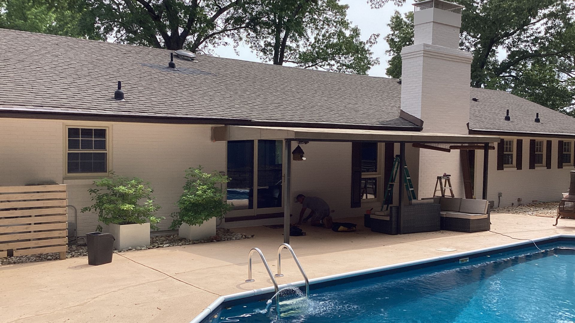 Man working on wood rot repair on a white brick house exterior with a pool