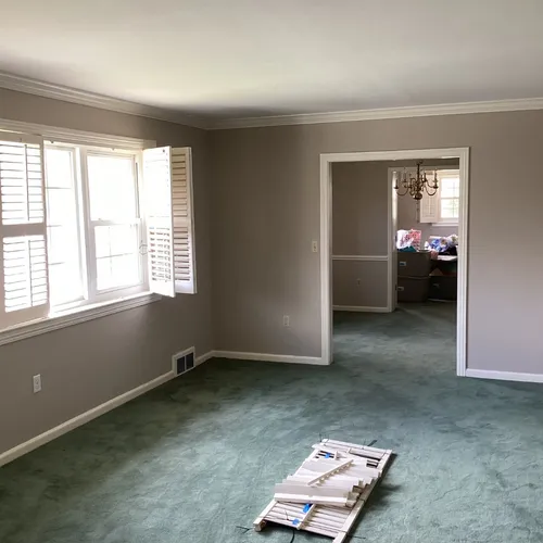 Empty living room with green carpet, white shutters, and doorway to dining room
